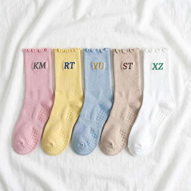 May include: Five pairs of colorful socks arranged on a white surface. Each sock is a different pastel color: pink, yellow, blue, tan, and white. Each sock has a different embroidered letter combination: KM, RT, YU, ST, and XZ.
