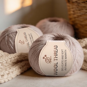May include: Three balls of light grey wool yarn are displayed, each with a label stating "WOOL THREAD, DELICATE AND SOFT, INTIMATE WARMTH." The yarn appears soft and fluffy. A cream-coloured knitted item is visible in the background, suitable for crafting.