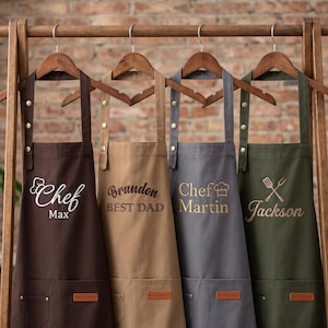 May include: Four personalized aprons in brown, tan, gray, and olive green hang on a wooden rack. Each apron features a different name and design: "Chef Max", "Brandon BEST DAD", "Chef Martin", and "Jackson" with a fork and spatula.