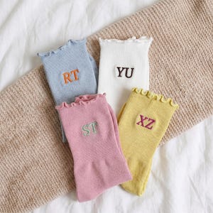 May include: Four pairs of colorful socks with ruffled tops and embroidered initials. The socks are in pastel shades of blue, white, pink, and yellow. Each sock has a different initial embroidered in a contrasting color.