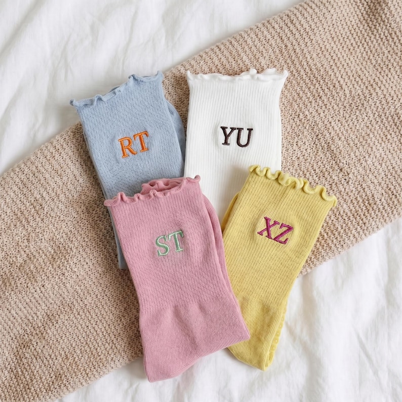 May include: Four pairs of colorful socks with ruffled tops and embroidered initials. The socks are in pastel shades of blue, white, pink, and yellow. Each sock has a different initial embroidered in a contrasting color.