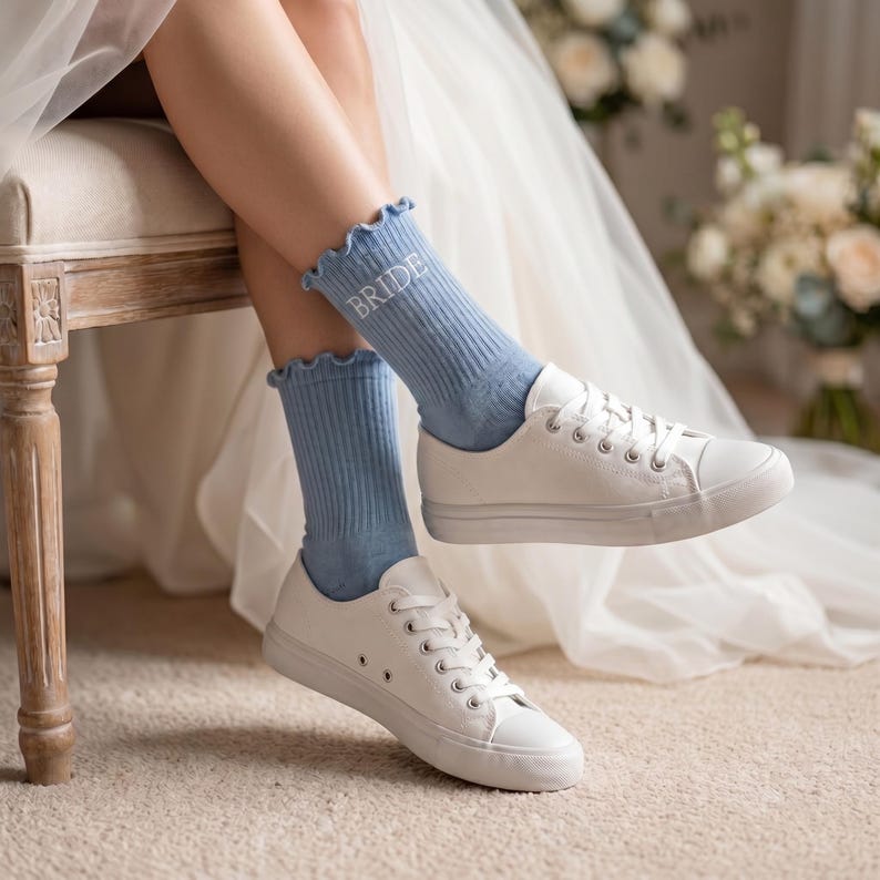May include: Light blue socks with the word "BRIDE" in white lettering, paired with white canvas sneakers. The socks have a ruffled edge. The scene is set indoors with a neutral-toned carpet and a wedding dress in the background.