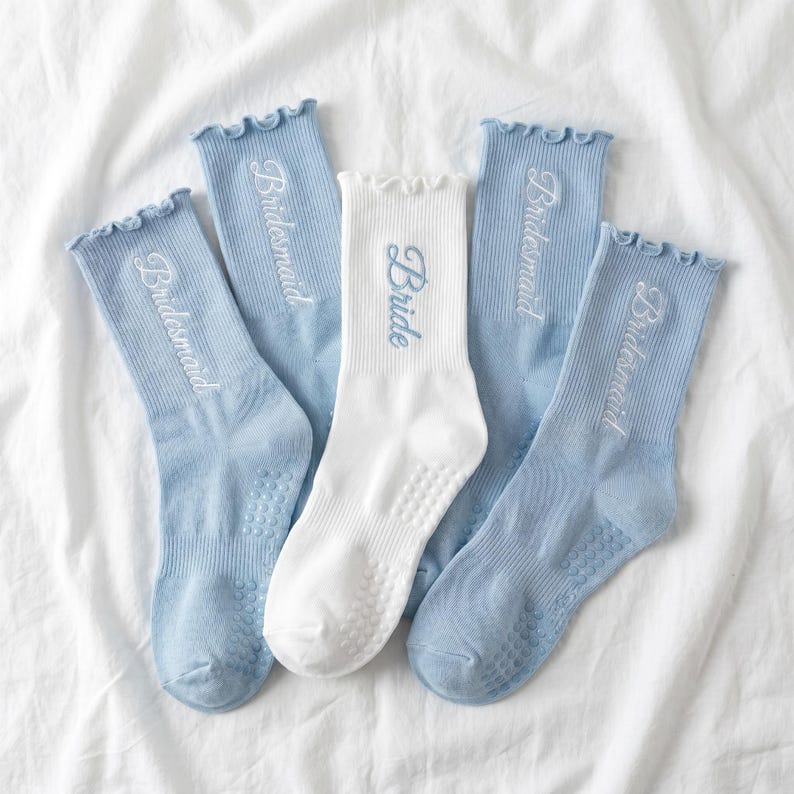 May include: Five pairs of socks, four light blue and one white, arranged on a white surface. The light blue socks have the word "Bridesmaid" embroidered in white, while the white sock says "Bride". All socks have a ruffled top.