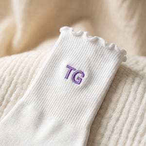 May include: Close-up of a white ribbed sock with a ruffled top edge. The sock features the embroidered letters "TG" in purple. The sock is made of a soft material and is likely for everyday wear.