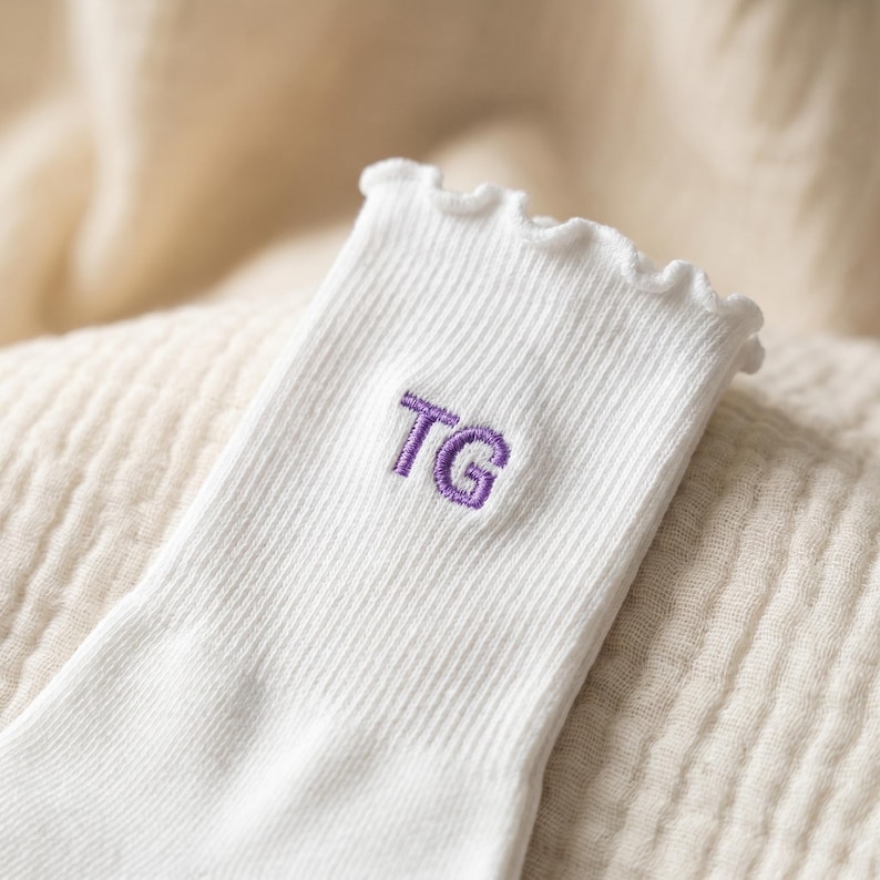 May include: Close-up of a white ribbed sock with a ruffled top edge. The sock features the embroidered letters "TG" in purple. The sock is made of a soft material and is likely for everyday wear.