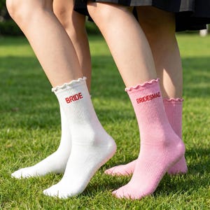 May include: White and pink crew socks with ruffled tops. The white sock has the word "BRIDE" in red, and the pink sock has "BRIDESMAID" in red. The socks are on a green grassy surface.