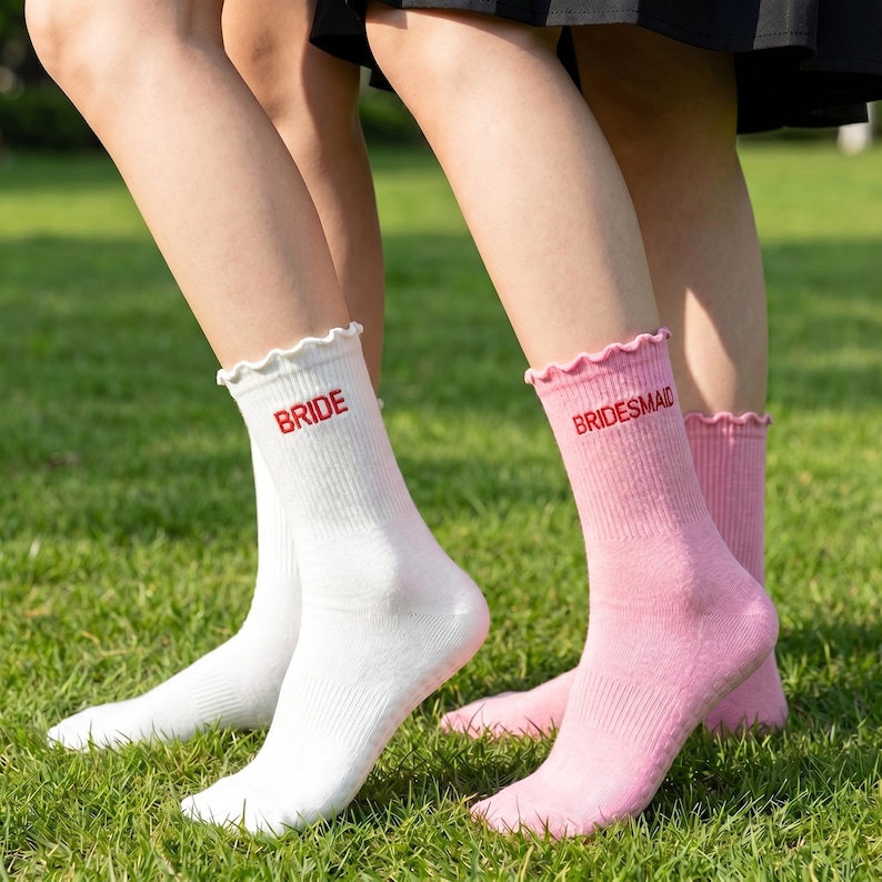 May include: White and pink crew socks with ruffled tops. The white sock has the word "BRIDE" in red, and the pink sock has "BRIDESMAID" in red. The socks are on a green grassy surface.