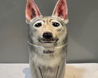 May include: A ceramic dog-shaped container with a lid, featuring a detailed painted face and upright ears. The container is primarily gray and white, with pink accents inside the ears. The name "Bailey" is written on the front.