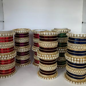May include: A collection of colorful bangles in red, purple, green, and blue. Each stack of bangles is adorned with gold-colored filigree and sparkling clear stones. The bangles are arranged in a row, showcasing their intricate designs.