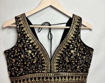 May include: Black sleeveless top with gold floral embroidery. The top has a deep V-neck and intricate gold detailing. The garment is displayed on a wooden hanger against a white backdrop.
