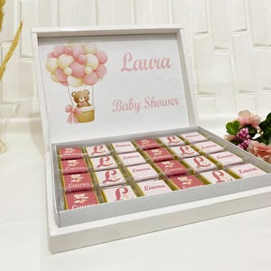 Puede incluir: Una caja blanca llena de bombones, ideal para un baby shower. La tapa presenta un diseño de globo aerostático rosa y blanco con un osito de peluche en una cesta, y el nombre "Laura" y "Baby Shower" en texto rosa.