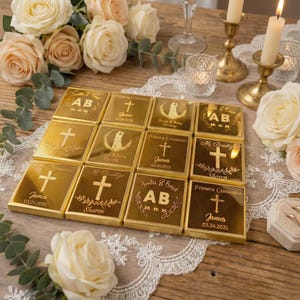May include: A collection of gold-colored, personalized favors, possibly for a wedding or baptism. Each square favor is engraved with names, dates, and religious symbols, such as crosses and wedding couple silhouettes. The favors are arranged on a table with floral and candle decorations.