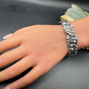 K&ouml;nnte beinhalten: Ein silberfarbenes Armband mit einem verzierten Blumenmuster. Das Armband weist detaillierte Schnitzereien von Blumen und Bl&auml;ttern auf, die eine Vintage-&Auml;sthetik erzeugen. Der Schmuck wird auf einer Mannequin-Hand pr&auml;sentiert.