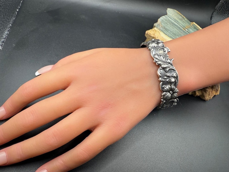 K&ouml;nnte beinhalten: Ein silberfarbenes Armband mit einem verzierten Blumenmuster. Das Armband weist detaillierte Schnitzereien von Blumen und Bl&auml;ttern auf, die eine Vintage-&Auml;sthetik erzeugen. Der Schmuck wird auf einer Mannequin-Hand pr&auml;sentiert.