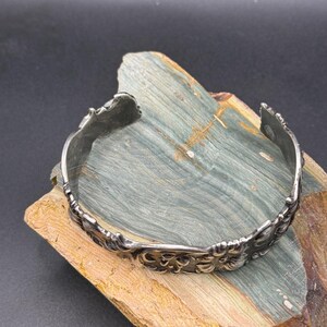 K&ouml;nnte beinhalten: Ein silberfarbenes Armreif-Armband mit einem verzierten floralen Design. Das offene Armband wird auf einem Holzst&uuml;ck mit Blau- und Braunt&ouml;nen pr&auml;sentiert. Die filigranen Details des Armbands sind sichtbar.