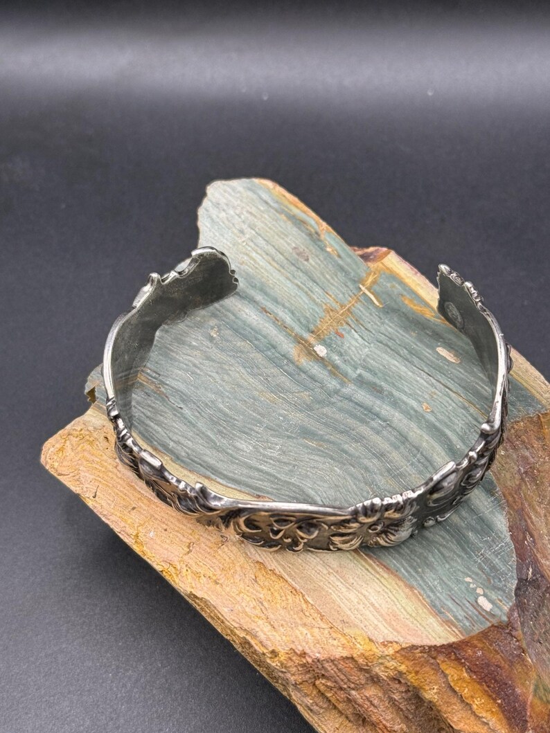 K&ouml;nnte beinhalten: Ein silberfarbenes Armreif-Armband mit einem verzierten floralen Design. Das offene Armband wird auf einem Holzst&uuml;ck mit Blau- und Braunt&ouml;nen pr&auml;sentiert. Die filigranen Details des Armbands sind sichtbar.
