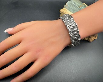 Könnte beinhalten: Ein silberfarbenes Armreif-Armband mit einem verzierten Blumenmuster. Das Armband ist breit und weist detaillierte Reliefschnitzereien von Blumen und Blättern auf. Das Armband wird an einem Handgelenk getragen.