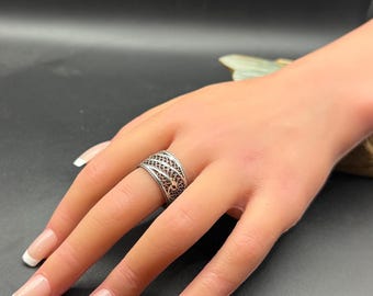 May include: A silver filigree ring with an openwork design. The ring is on a hand with a French manicure. The ring's intricate pattern is visible, showcasing a detailed, open weave. The ring is a piece of jewellery.