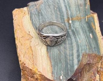 Könnte beinhalten: Silberner Ring mit ägyptischem Skarabäus-Design. Der Ring zeigt ein detailliertes Skarabäus-Emblem mit einem kleinen, runden, roten Stein in der Mitte. Der Ring ist vor dem Hintergrund eines farbenfrohen, strukturierten Steins platziert.