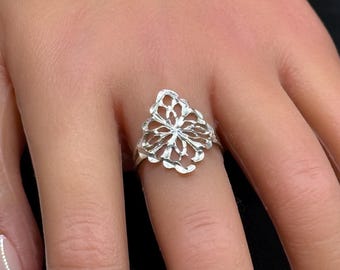 May include: A silver ring with an openwork floral design. The ring's intricate pattern resembles a stylized flower or leaf. The ring is worn on a finger, showcasing its elegant and airy aesthetic. The ring's design is detailed and intricate, with multiple petals.
