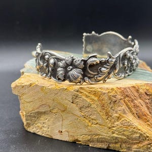 K&ouml;nnte beinhalten: Ein silberfarbenes Armreif-Armband mit aufwendigen Blumen- und Blattdetails. Das offene Armband weist ein detailliertes Design aus Blumen und Bl&auml;ttern auf und erzeugt eine Vintage- oder Antik-&Auml;sthetik. Die Metallarbeiten sind sehr detailliert und zeigen die Handwerkskunst.