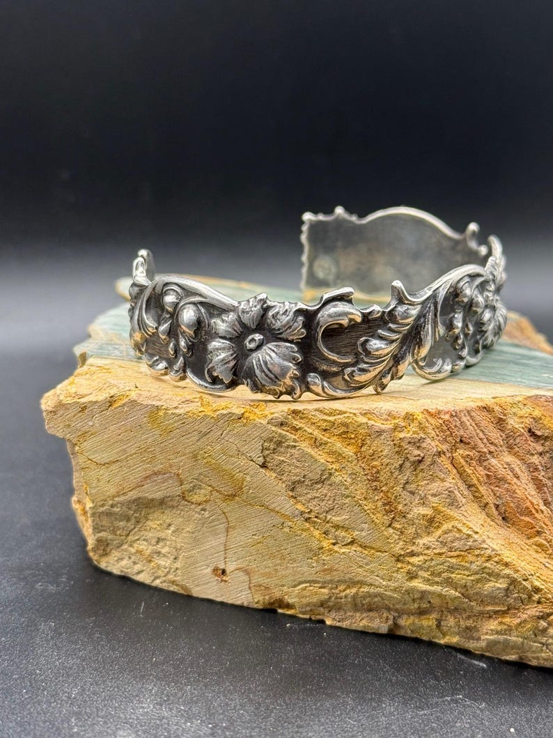 K&ouml;nnte beinhalten: Ein silberfarbenes Armreif-Armband mit aufwendigen Blumen- und Blattdetails. Das offene Armband weist ein detailliertes Design aus Blumen und Bl&auml;ttern auf und erzeugt eine Vintage- oder Antik-&Auml;sthetik. Die Metallarbeiten sind sehr detailliert und zeigen die Handwerkskunst.