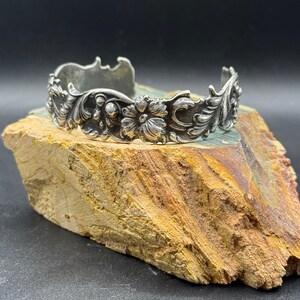 K&ouml;nnte beinhalten: Ein silberfarbenes Armreif-Armband mit einem verzierten Blumen- und Blattdesign. Das Armband weist detaillierte Schnitzereien von Blumen und Bl&auml;ttern auf, die eine Vintage-&Auml;sthetik erzeugen. Der Armreif wird auf einer Natursteinoberfl&auml;che pr&auml;sentiert.