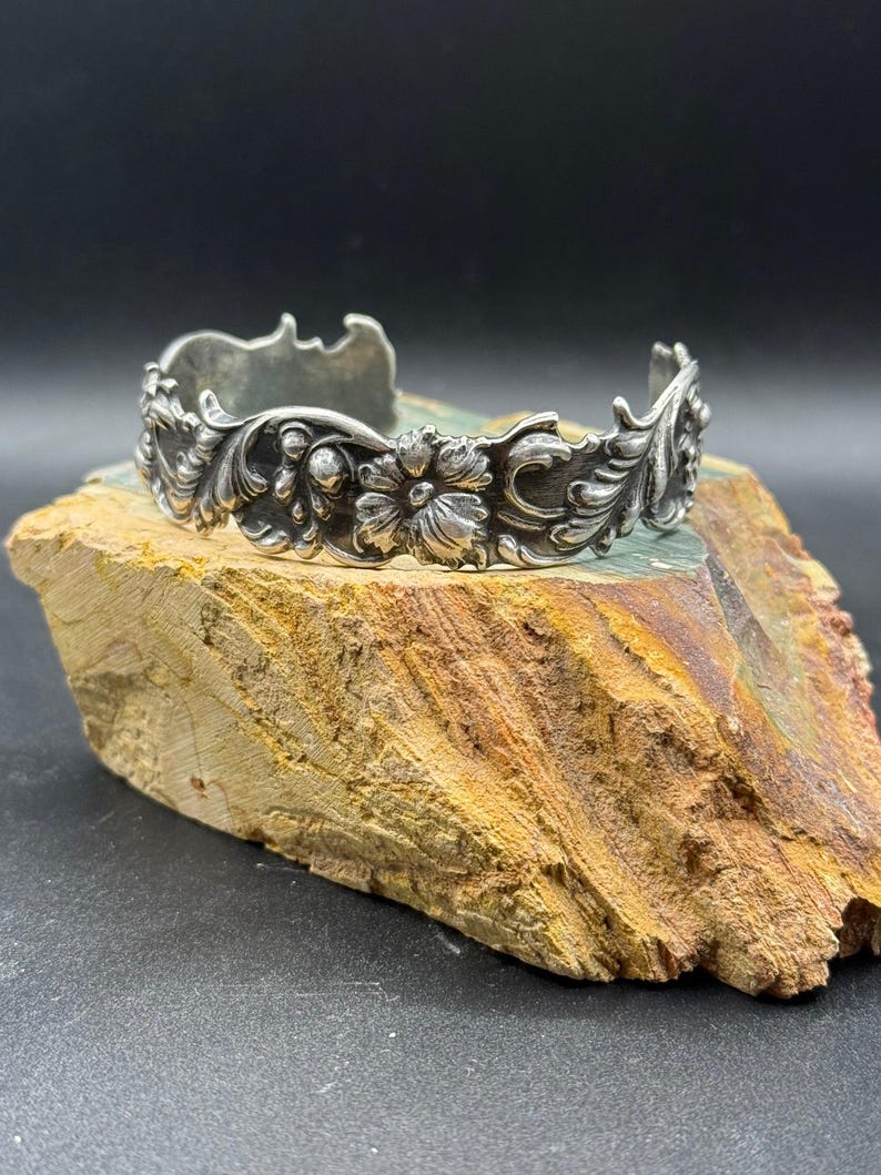K&ouml;nnte beinhalten: Ein silberfarbenes Armreif-Armband mit einem verzierten Blumen- und Blattdesign. Das Armband weist detaillierte Schnitzereien von Blumen und Bl&auml;ttern auf, die eine Vintage-&Auml;sthetik erzeugen. Der Armreif wird auf einer Natursteinoberfl&auml;che pr&auml;sentiert.