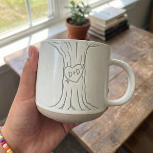 Puede incluir: Taza de cerámica blanca con un diseño de árbol y un corazón con "D+D" grabado. La taza tiene una base marrón claro y un asa grande. La taza se sostiene en una mano.