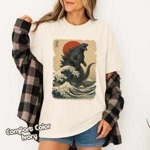 May include: An off-white t-shirt featuring a graphic print of a dark green monster standing in front of a large red sun and a stylized ocean wave. The design includes a small vertical rectangular box with Japanese text in black ink.