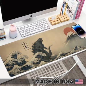 May include: A long rectangular desk mat featuring a Godzilla design inspired by Japanese art. The design includes Godzilla, Mount Fuji, and ocean waves in shades of grey, blue, and beige. The text "MADE IN USA" is at the bottom.