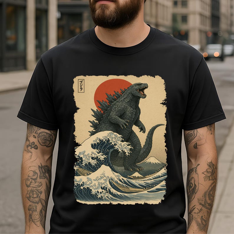 May include: A black t-shirt featuring a graphic print of a dark grey Godzilla standing in front of a large red sun. The design includes stylized blue ocean waves, a mountain in the background, and the Japanese text ゴジラ in a small white box.