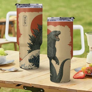 May include: Two stainless steel travel mugs with white handles. Each mug features a Godzilla design with a red sun, Japanese text, and a wave pattern. The mugs are on a wooden table.