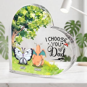 May include: A heart-shaped clear acrylic plaque featuring a printed illustration of a butterfly and a dragon-like creature in a grassy field under a tree. The text reads I CHOOSE YOU Dad with a small Poké Ball icon, ROSA, and DADDY printed below.