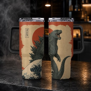 May include: A stainless steel tumbler with a black handle and clear lid, featuring a graphic of Godzilla against a red sun and stylized ocean waves. The design includes the Japanese text ゴジラ in a vertical rectangular box on a beige background.