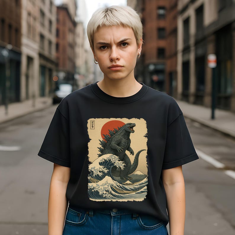 May include: A black short-sleeved t-shirt featuring a graphic print of a dark grey monster standing amidst stylized ocean waves. The design includes a large red sun in the background, a mountain silhouette, and Japanese characters in a rectangular border.
