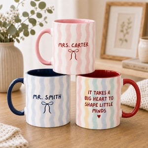 May include: Three ceramic mugs with wavy vertical stripes. The top mug is pink and white with 'MRS. CARTER' and a bow. The bottom left is blue and white with 'MR. SMITH' and a bow. The bottom right is multicoloured with 'IT TAKES A BIG HEART TO SHAPE LITTLE MINDS'.