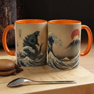 Könnte beinhalten: Zwei Keramikbecher mit orangefarbenen Griffen und Innenräumen. Ein Becher zeigt Godzilla in einer Welle, der andere einen Berg und eine Sonne. Die Becher haben einen beigefarbenen Hintergrund im japanischen Kunststil.