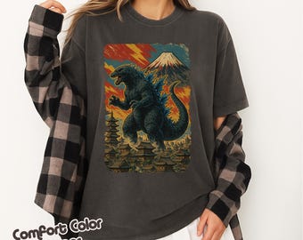 May include: Dark gray t-shirt featuring a Godzilla graphic. The graphic depicts Godzilla in front of a mountain and buildings, with a colorful sky. The text "Comfort Color Pepper" is at the bottom left.