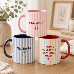 May include: Three ceramic mugs with vertical stripes. The top mug is pink and white with the text Mrs. Carter and a bow. The bottom left mug is blue and white with the text Mr. Smith and a bow. The bottom right mug has multicolored stripes and the text It takes a big heart to shape little minds with a heart icon.