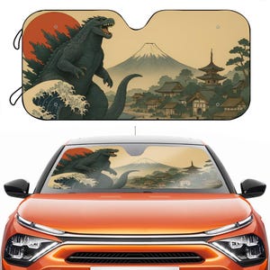 May include: Car sunshade featuring a Godzilla design with a Japanese landscape backdrop. The artwork includes a red sun, a mountain, and traditional buildings. The sunshade is black with a cutout for the rearview mirror and two black loops.