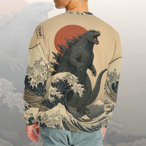 Può includere: Felpa beige con un design di Godzilla, un sole rosso, onde oceaniche e una montagna. Il testo giapponese "ゴジラ" è in un riquadro. L'opera d'arte è in stile Ukiyo-e giapponese.