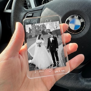 Peut inclure: Support photo en acrylique transparent avec une photo de mariage en noir et blanc. La photo montre des mariés se tenant la main devant un château. Le texte "Driver Safe" et "I Love You" sont écrits sur le support, avec les noms "John & Julia".