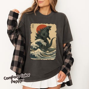 May include: A charcoal gray t-shirt featuring a graphic print of a green monster standing in a large ocean wave with a red sun and Mount Fuji in the background. The design includes Japanese text in a vertical box. A black and gray plaid shirt is worn over it.