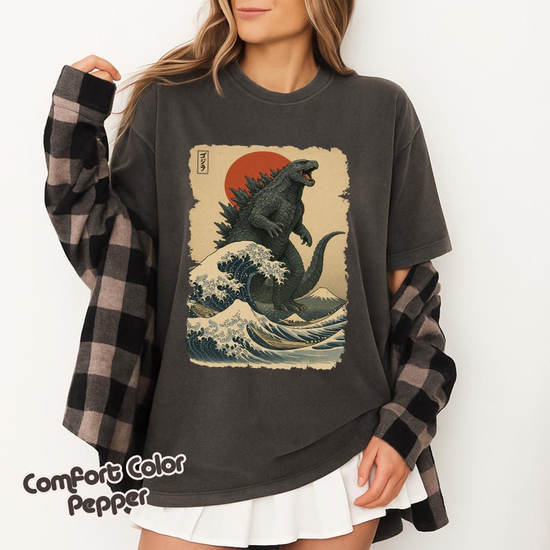 May include: A charcoal gray t-shirt featuring a graphic print of a green monster standing in a large ocean wave with a red sun and Mount Fuji in the background. The design includes Japanese text in a vertical box. A black and gray plaid shirt is worn over it.