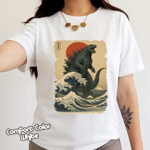 May include: A white short-sleeved t-shirt featuring a graphic print of a dark grey Godzilla standing in front of a large red sun. The design includes a stylized blue ocean wave, a mountain in the background, and Japanese text in a small box.