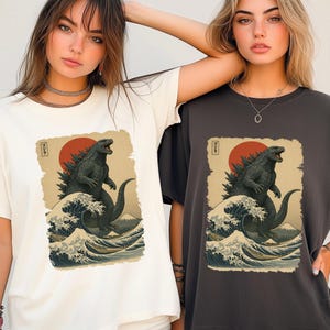 May include: A white t-shirt and a dark gray t-shirt, each featuring a graphic print of a dark green monster standing in ocean waves with a large red sun and a mountain in the background. The print includes Japanese text in a small vertical box.
