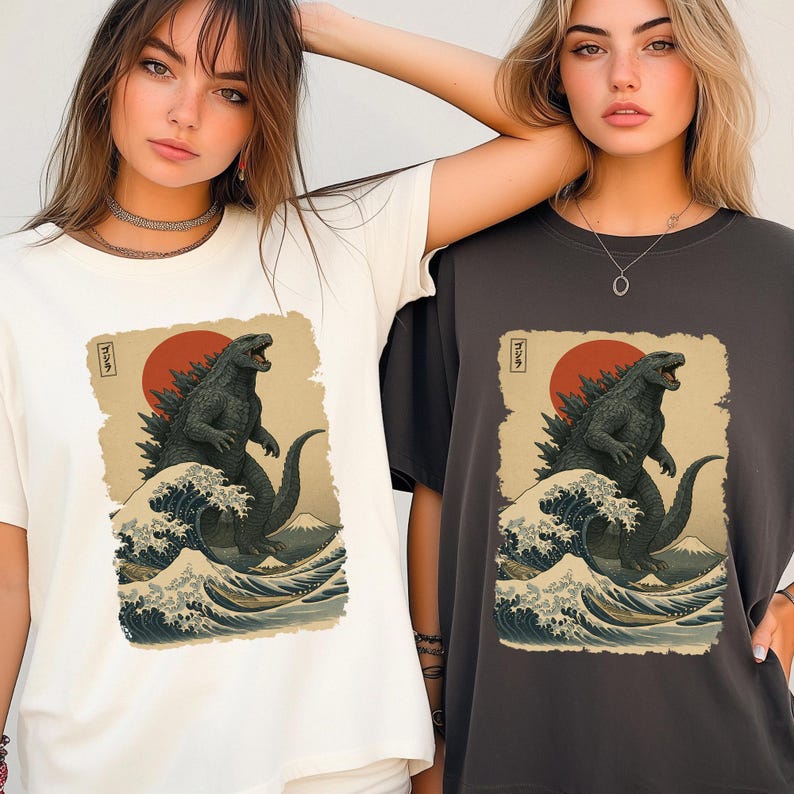 May include: A white t-shirt and a dark gray t-shirt, each featuring a graphic print of a dark green monster standing in ocean waves with a large red sun and a mountain in the background. The print includes Japanese text in a small vertical box.