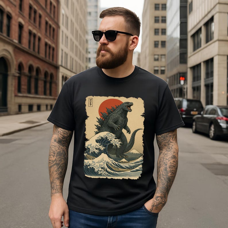 May include: A black t-shirt featuring a graphic print of a large monster standing amidst stylized ocean waves, a red sun, and a mountain in the background. The design includes Japanese text in the upper left corner, set against a cream-colored rectangular background.