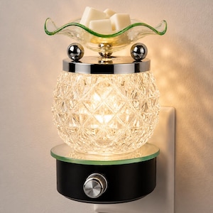 May include: A plug-in wax warmer featuring a clear, diamond-patterned glass body with a chrome top and two small silver spheres. A wavy-edged glass dish sits on top, holding several white wax cubes. The base is black with a silver control knob.